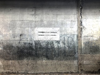 A textured, worn concrete wall with visible cracks and stains, featuring a sign in Spanish that prohibits the posting of posters. The surface appears aged and industrial.