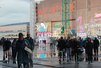 A diverse group of people walking across an urban space with reflective puddles on the ground. The background features a large wall with colorful graffiti and text, including a visible phrase 'MAKE ART' in bold red lettering. A green construction crane is also present, indicating ongoing development. The atmosphere is bustling and dynamic, with people dressed in casual clothing for various weather conditions.