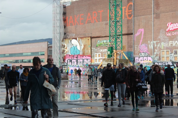 A diverse group of people walking across an urban space with reflective puddles on the ground. The background features a large wall with colorful graffiti and text, including a visible phrase 'MAKE ART' in bold red lettering. A green construction crane is also present, indicating ongoing development. The atmosphere is bustling and dynamic, with people dressed in casual clothing for various weather conditions.