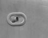 A metal surface with an oval-shaped peephole, through which a protruding nozzle or pipe is visible. The entire image is in shades of gray, reflecting an industrial or mechanical theme.