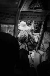 Two people are inside a vehicle. One person is wearing a large white hat and a striped shirt, holding a bag. The other person, dressed in a uniform, is assisting or interacting with them. Signage with instructions is visible in the vehicle.