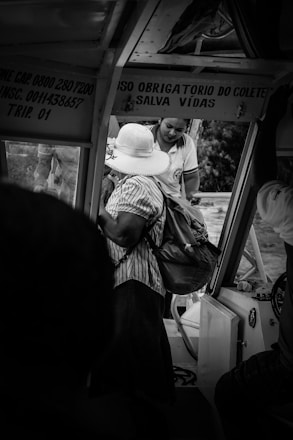 Two people are inside a vehicle. One person is wearing a large white hat and a striped shirt, holding a bag. The other person, dressed in a uniform, is assisting or interacting with them. Signage with instructions is visible in the vehicle.