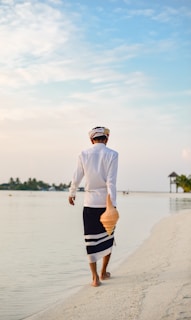 A person dressed in traditional clothing walks along a tranquil beach. They hold a large conch shell in one hand while barefoot on the sandy shore. The sky is clear with soft clouds, and the horizon features a glimpse of a small pavilion and lush greenery on the side.