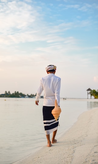 A person dressed in traditional clothing walks along a tranquil beach. They hold a large conch shell in one hand while barefoot on the sandy shore. The sky is clear with soft clouds, and the horizon features a glimpse of a small pavilion and lush greenery on the side.