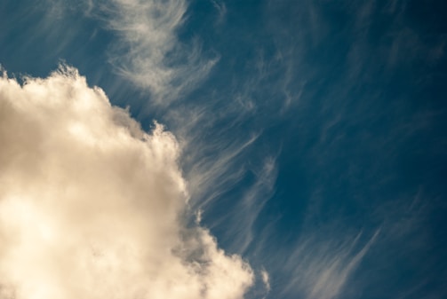 A large, fluffy cloud occupies the left side of the image, with delicate wisps of cloud stretching out over a deep blue sky. The lighting creates a warm glow on the clouds, enhancing the contrast against the cool background.