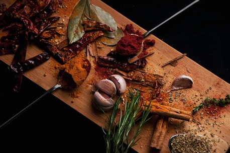 A wooden cutting board covered with a variety of spices including whole dried red chilies, bay leaves, a garlic bulb, rosemary sprigs, cinnamon sticks, and ground spices scattered around. Two metal spoons hold rich red and orange powders. There is also a small pile of green herbs on the board.