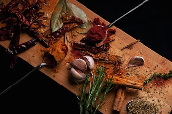 A wooden cutting board covered with a variety of spices including whole dried red chilies, bay leaves, a garlic bulb, rosemary sprigs, cinnamon sticks, and ground spices scattered around. Two metal spoons hold rich red and orange powders. There is also a small pile of green herbs on the board.
