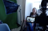 A camera is set up for recording, with a person visible on the camera's display screen. The person appears to be in a sitting position, possibly being interviewed or giving a talk. A green backdrop and additional lighting equipment suggest a professional or semi-professional filming setting.