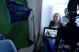 A camera is set up for recording, with a person visible on the camera's display screen. The person appears to be in a sitting position, possibly being interviewed or giving a talk. A green backdrop and additional lighting equipment suggest a professional or semi-professional filming setting.