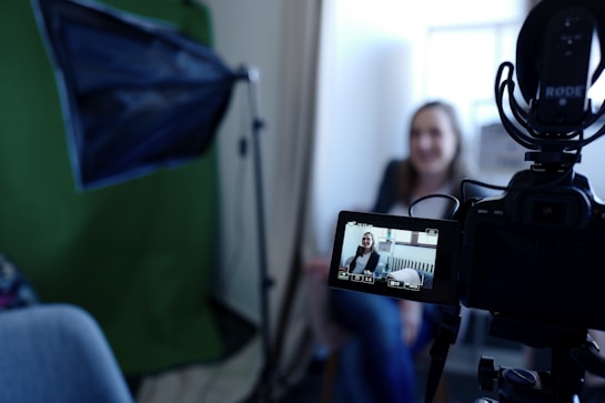 A camera is set up for recording, with a person visible on the camera's display screen. The person appears to be in a sitting position, possibly being interviewed or giving a talk. A green backdrop and additional lighting equipment suggest a professional or semi-professional filming setting.