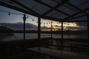 Outdoor seating area with wooden benches and greenery beside the water at sunset.