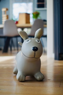 A playful grey dog sculpture sits on a wooden floor in a cozy room. The background includes blurred chairs and a table, with plants and decor adding warmth to the space.