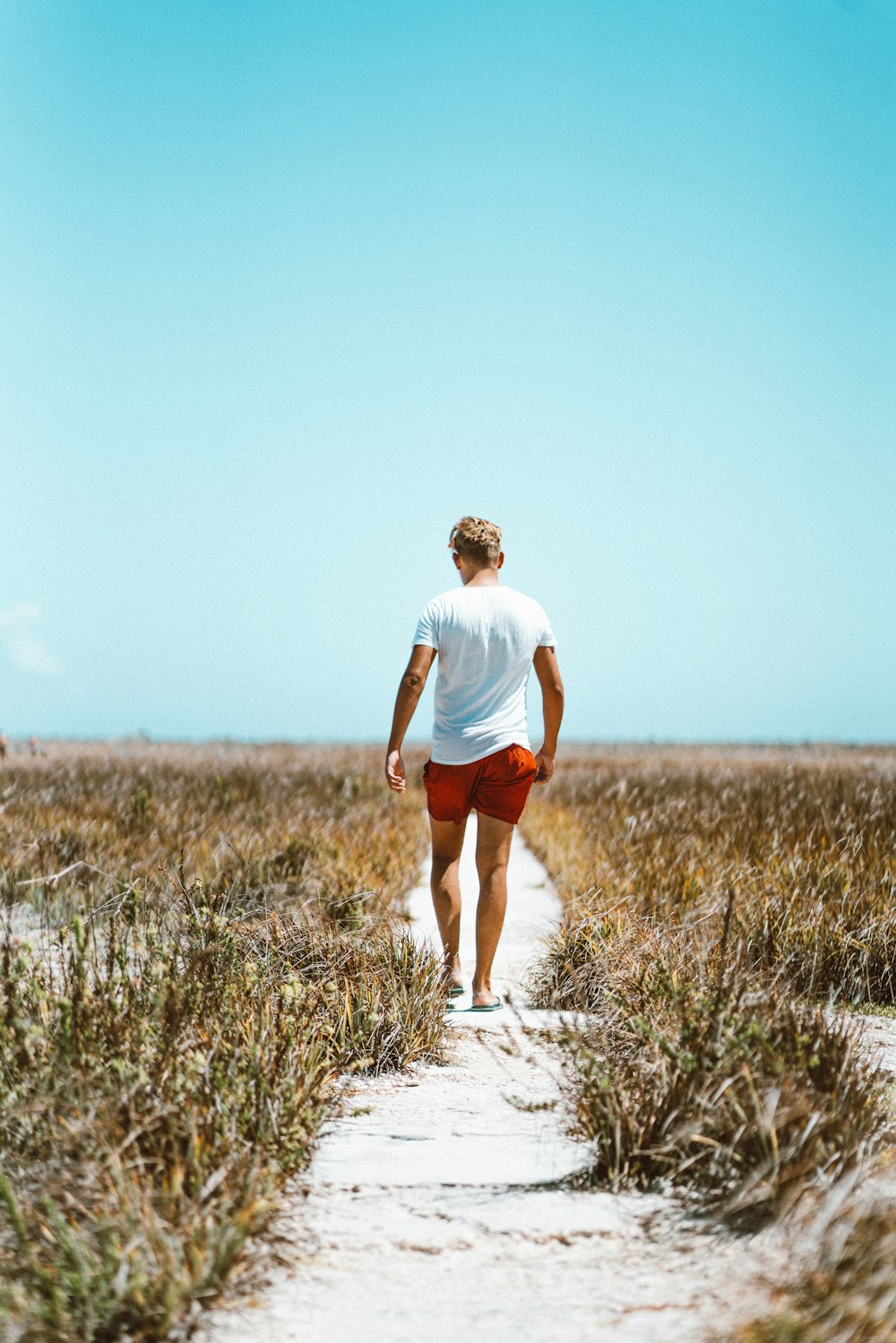 草むらの間を白い砂の上を歩く白いシャツと赤い短パンの男性