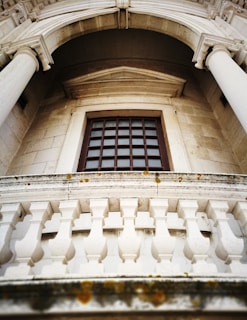 An upward view of a classical architectural facade featuring tall stone columns and a window with a grid pattern. A decorative stone balustrade is visible in the foreground, with some moss growing on the stone. The stonework exhibits intricate detailing and craftsmanship, characteristic of historic structures.