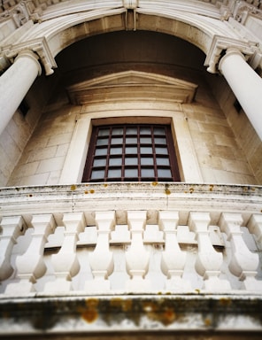 An upward view of a classical architectural facade featuring tall stone columns and a window with a grid pattern. A decorative stone balustrade is visible in the foreground, with some moss growing on the stone. The stonework exhibits intricate detailing and craftsmanship, characteristic of historic structures.