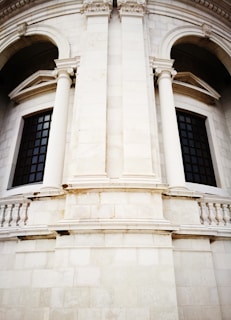 A detailed view of a classical architectural structure featuring tall, white stone columns flanking large, arched windows with black grilles. The building facade includes ornate detailing around the columns and arches, creating a sense of grandeur.