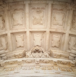 An intricately designed architectural ceiling with ornate details and patterns. The surface features symmetrical, recessed panels framed by a series of moldings. Below, a decorative arch contains artistic carvings, including figures and floral elements.