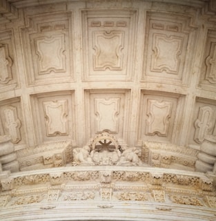 An intricately designed architectural ceiling with ornate details and patterns. The surface features symmetrical, recessed panels framed by a series of moldings. Below, a decorative arch contains artistic carvings, including figures and floral elements.