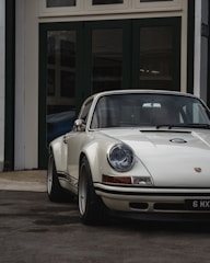 A classic white Porsche car with a vintage design is parked in front of dark green garage doors. The car's round headlights and sleek body lines are prominent, reflecting a timeless, elegant style.