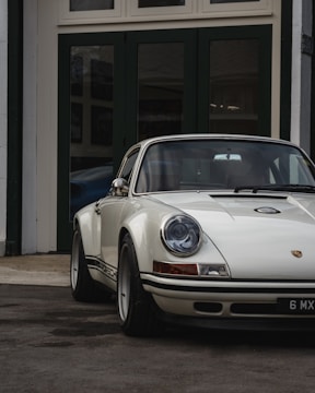 A classic white Porsche car with a vintage design is parked in front of dark green garage doors. The car's round headlights and sleek body lines are prominent, reflecting a timeless, elegant style.