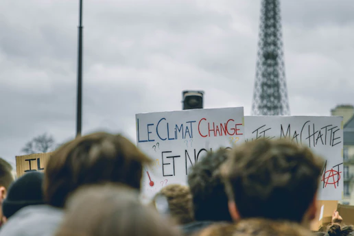 A protest gathering with signs held by participants. One prominent sign reads 'Le Climat Change,' indicating a focus on climate change. The Eiffel Tower is visible in the background, suggesting a location in Paris. The atmosphere appears crowded with a diverse group of people.