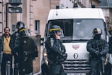 A group of riot police officers in protective gear stand near a police van. They are equipped with helmets, shields, and other protective equipment. One officer is speaking on a radio. A man in civilian clothing is visible in the background, watching the scene.