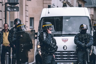 A group of riot police officers in protective gear stand near a police van. They are equipped with helmets, shields, and other protective equipment. One officer is speaking on a radio. A man in civilian clothing is visible in the background, watching the scene.