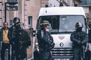 A group of riot police officers in protective gear stand near a police van. They are equipped with helmets, shields, and other protective equipment. One officer is speaking on a radio. A man in civilian clothing is visible in the background, watching the scene.