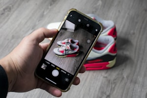 A hand holds a smartphone capturing a photo of a pair of sneakers placed on a wooden floor. The sneakers have a red, white, and black color scheme with visible branding details.