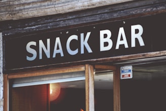 A sign with bold white letters spells out 'SNACK BAR' against a dark background. Below the sign, a partial view of a glass door is visible with a small sign indicating that it is currently closed. The surrounding area shows aged wooden and stone elements, giving it a rustic look.