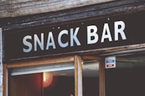 A sign with bold white letters spells out 'SNACK BAR' against a dark background. Below the sign, a partial view of a glass door is visible with a small sign indicating that it is currently closed. The surrounding area shows aged wooden and stone elements, giving it a rustic look.