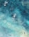 Bird's-eye view of people surfing or paddling in the ocean. The water appears clear with varying shades of blue and turquoise, and sunlight reflects off the surface. There are three individuals visible, each on a surfboard, separated by the vast, shimmering water.