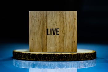 A wooden block with the word 'LIVE' printed on it is placed on a circular wooden slice. The setup is against a dark background with a reflective surface underneath, creating a mirror-like effect.