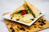 A white square plate holds a sandwich with waffle-textured bread filled with lettuce, green vegetables, and possibly some protein. Accompanying the sandwich are potato chips seasoned with herbs and a whole strawberry. The plate is placed on a wooden surface, surrounded by an assortment of nuts and raisins.