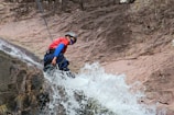A person wearing a helmet and protective gear rappels down a rocky cliff with water cascading over the rocks. The individual is equipped with ropes and is wearing a red shirt and sunglasses. Water splashes energetically in the foreground, adding a dynamic element.