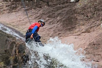 A person wearing a helmet and protective gear rappels down a rocky cliff with water cascading over the rocks. The individual is equipped with ropes and is wearing a red shirt and sunglasses. Water splashes energetically in the foreground, adding a dynamic element.