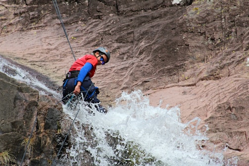 A person wearing a helmet and protective gear rappels down a rocky cliff with water cascading over the rocks. The individual is equipped with ropes and is wearing a red shirt and sunglasses. Water splashes energetically in the foreground, adding a dynamic element.