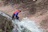 A smiling woman wearing a helmet and harness, standing at the edge of a cliff, ready to rappel.
