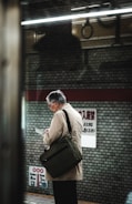 An elderly man with gray hair stands on a subway platform, reading a document or device. He is wearing a light-colored coat and carrying a dark shoulder bag. The background includes a brick-patterned wall and signage with text in a non-Latin script. The lighting is somewhat dim, suggesting an indoor setting with artificial lights.