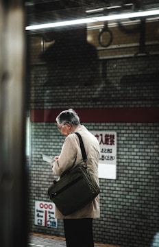 An elderly man with gray hair stands on a subway platform, reading a document or device. He is wearing a light-colored coat and carrying a dark shoulder bag. The background includes a brick-patterned wall and signage with text in a non-Latin script. The lighting is somewhat dim, suggesting an indoor setting with artificial lights.