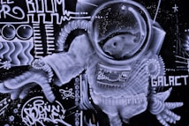 A detailed graffiti artwork featuring an astronaut in a spacesuit, set against a backdrop of abstract designs and patterns. The astronaut's helmet is prominent, adorned with intricate linework, while the suit is decorated with various symbols, including numbers and geometric shapes. The word 'GALACT' is visible, accompanied by other abstract figures and artistic elements that give a sense of movement and energy.
