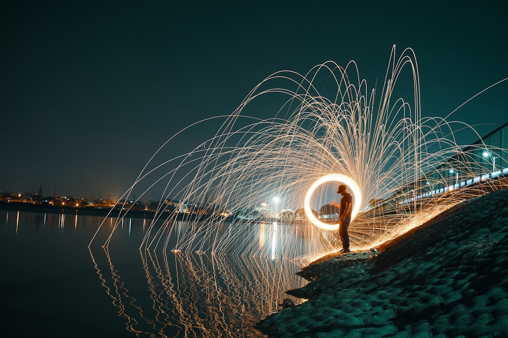 time lapse photography of man spinning lighted steel wool near body of water