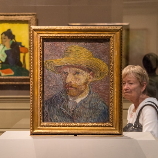 A framed painting of a man wearing a straw hat sits on display in a museum. The painting features impressionistic brushwork and vibrant colors. Surrounding the painting are other artworks and a visitor observing the display.
