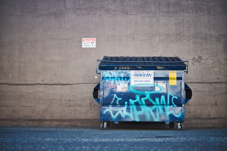 A large, blue metal dumpster with graffiti is positioned against a concrete wall. The wall has a small, red and white warning sign about security cameras. The dumpster features various stickers and notices, and its lid is partially opened. The ground appears to be a flat, asphalt surface.