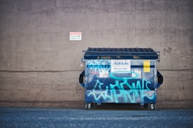 A large, blue metal dumpster with graffiti is positioned against a concrete wall. The wall has a small, red and white warning sign about security cameras. The dumpster features various stickers and notices, and its lid is partially opened. The ground appears to be a flat, asphalt surface.