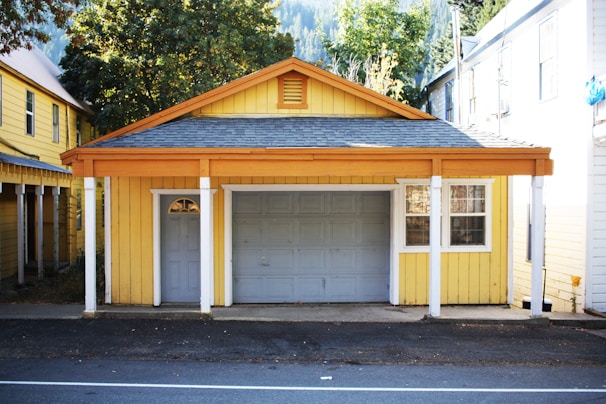 A small yellow garage with white pillars and a gray roof is situated along a paved road. It has a closed gray garage door and a small window to the right. Surrounding the garage are additional yellow and white buildings, as well as trees and a partially visible mountain landscape in the background.