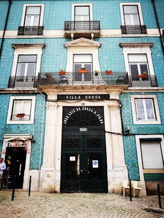 A historic building facade featuring a large, ornate black door with the inscription 'VILLA SOUSA' above and the date '1890'. The building has multiple windows with white frames, some adorned with small balconies and potted plants. The walls are covered in turquoise tiles, adding a unique texture. There is a small shop entrance on the left and a couple of chairs outside on the right.