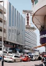 A busy urban street scene features tall buildings with modern architecture, including a tall glass-fronted hotel. Numerous cars, including red and white vehicles, are parked and moving along the street. A prominent sign reading 'Al Madina Supermarket' is visible, and a person is seen rappelling down one of the building facades.
