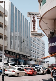 A busy urban street scene features tall buildings with modern architecture, including a tall glass-fronted hotel. Numerous cars, including red and white vehicles, are parked and moving along the street. A prominent sign reading 'Al Madina Supermarket' is visible, and a person is seen rappelling down one of the building facades.