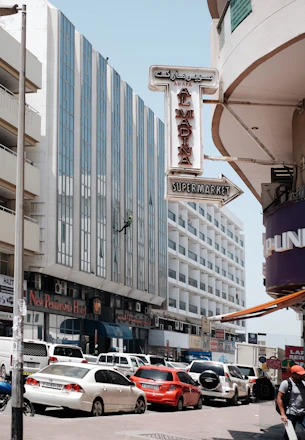 A busy urban street scene features tall buildings with modern architecture, including a tall glass-fronted hotel. Numerous cars, including red and white vehicles, are parked and moving along the street. A prominent sign reading 'Al Madina Supermarket' is visible, and a person is seen rappelling down one of the building facades.
